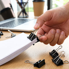 A hand holds a black binder clip on a stack of white paper, with a laptop and other clips in the background.