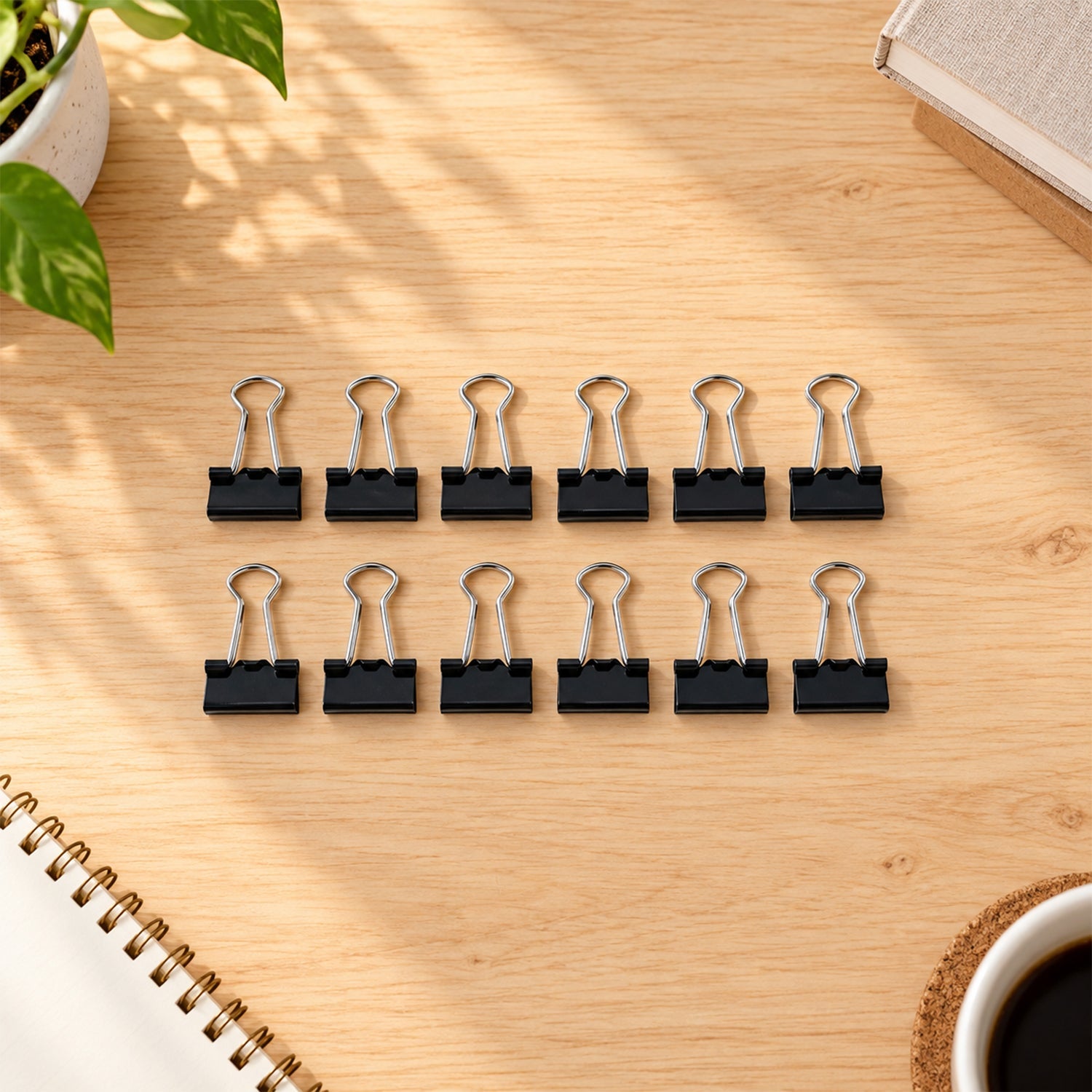 Twelve black binder clips with silver handles are arranged on a light wooden desk, next to a notebook and a plant.