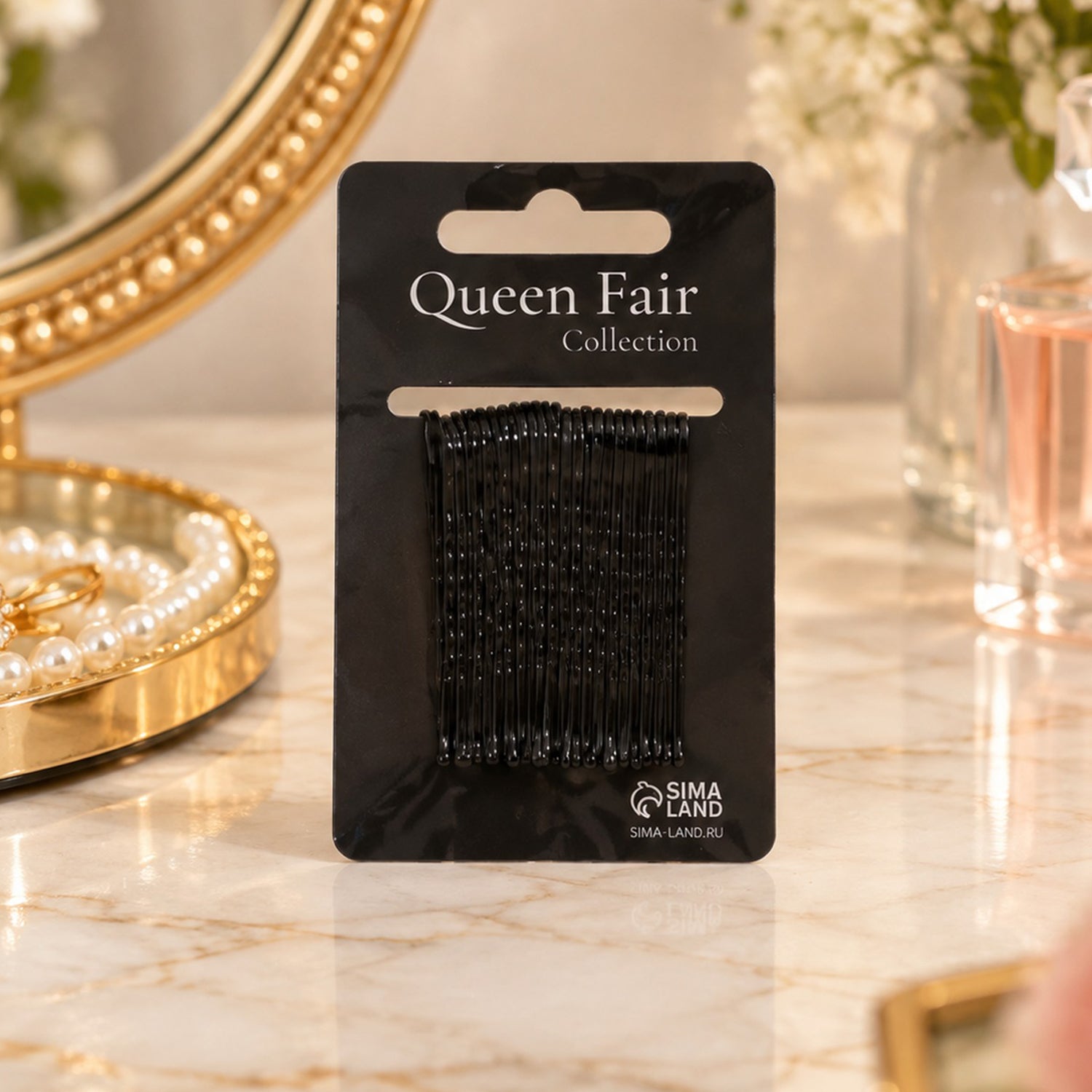 A black card of bobby pins stands on a marble surface next to a gold mirror and pearl necklace.