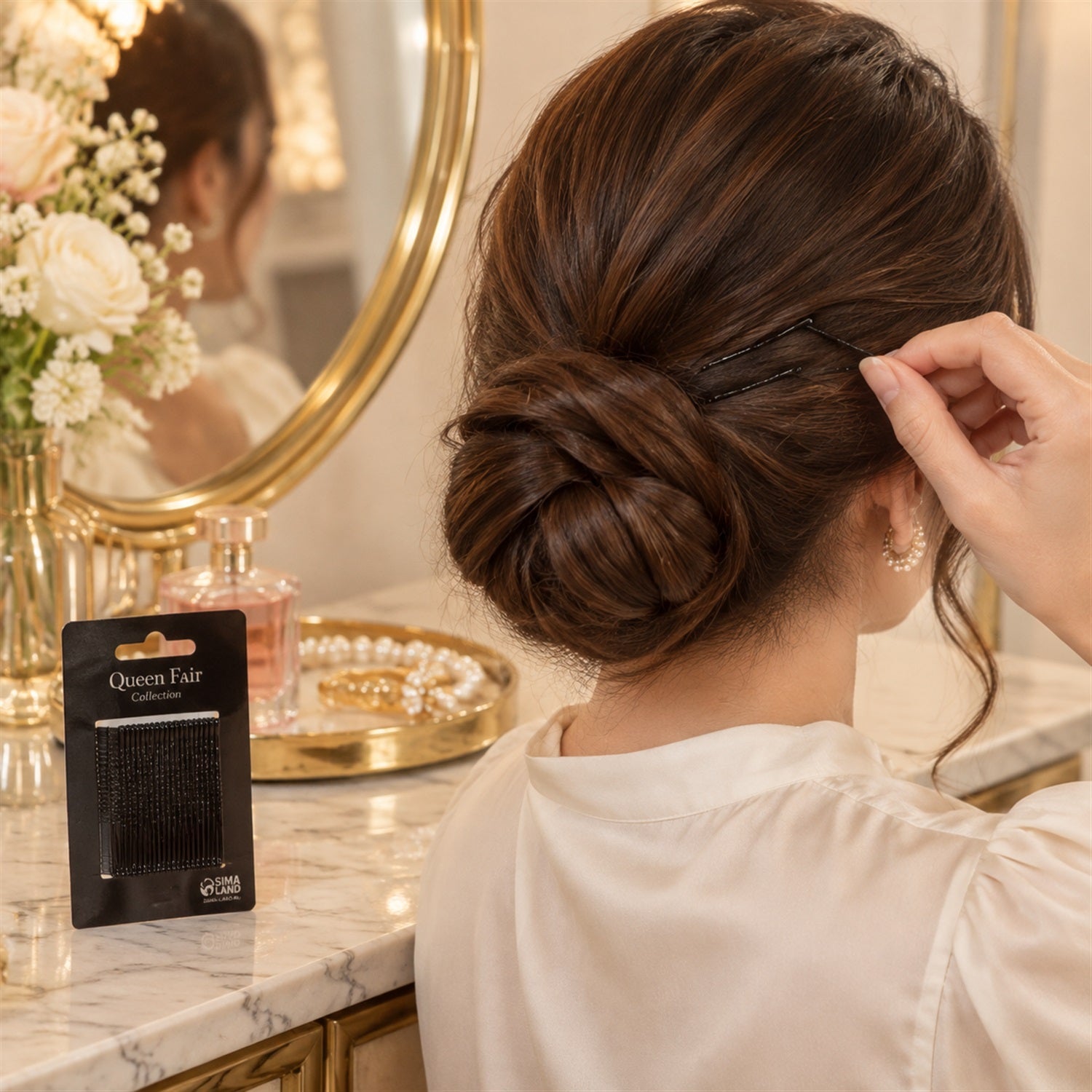 A woman uses a black bobby pin to style her hair bun, with a mirror and perfume bottle in the background.