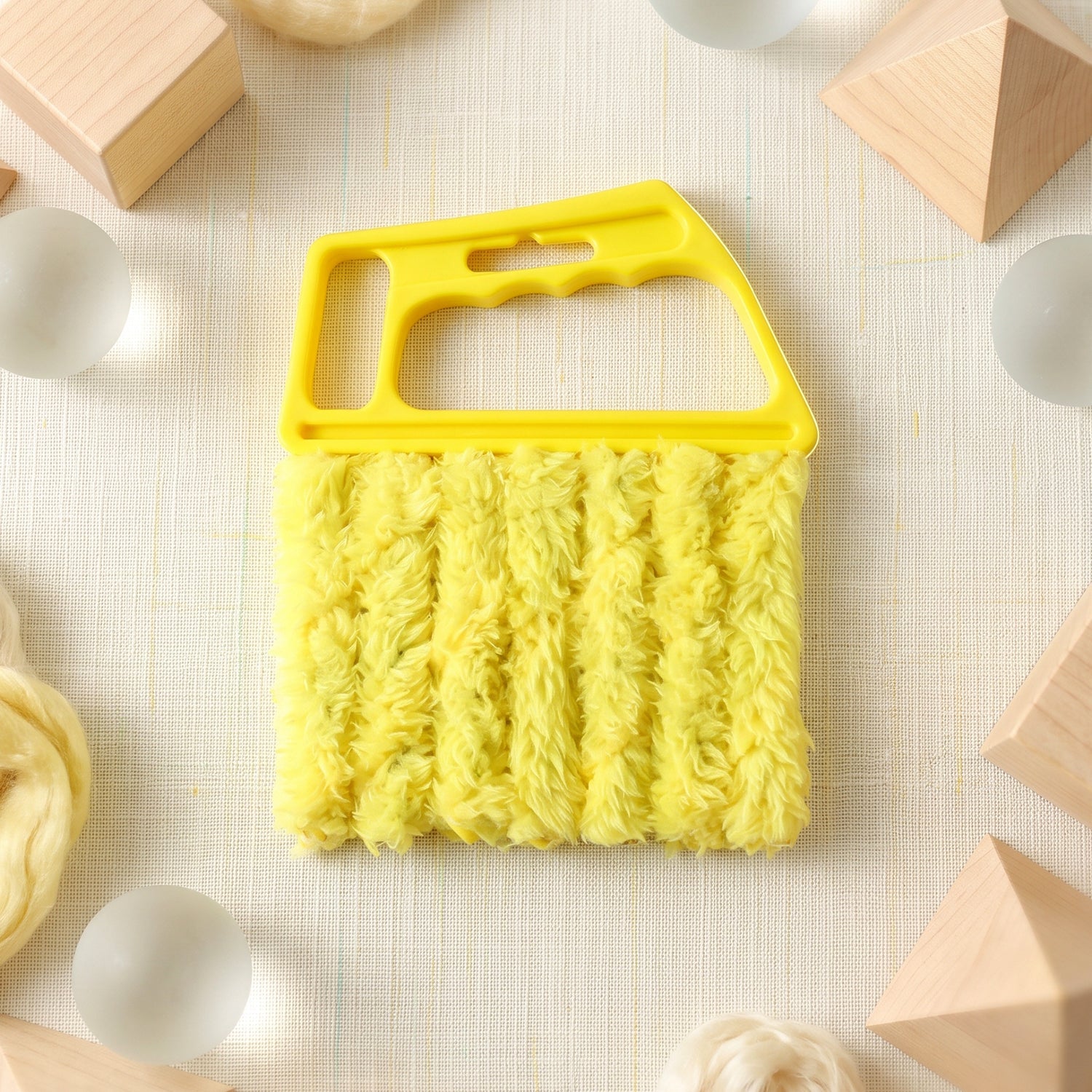 A yellow microfiber blind cleaner is centered on a textured white surface, surrounded by wooden blocks and glass spheres.