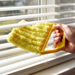 A hand holds a bright yellow microfiber blind cleaner, wiping dust from white horizontal window blinds.