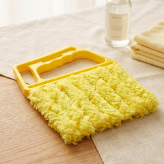 A yellow microfiber blind cleaner rests on a light tablecloth, with a spray bottle and folded yellow cloths nearby.
