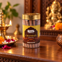 A transparent jar of brown dhoop sticks sits on a wooden surface with a lit lamp and flowers nearby.