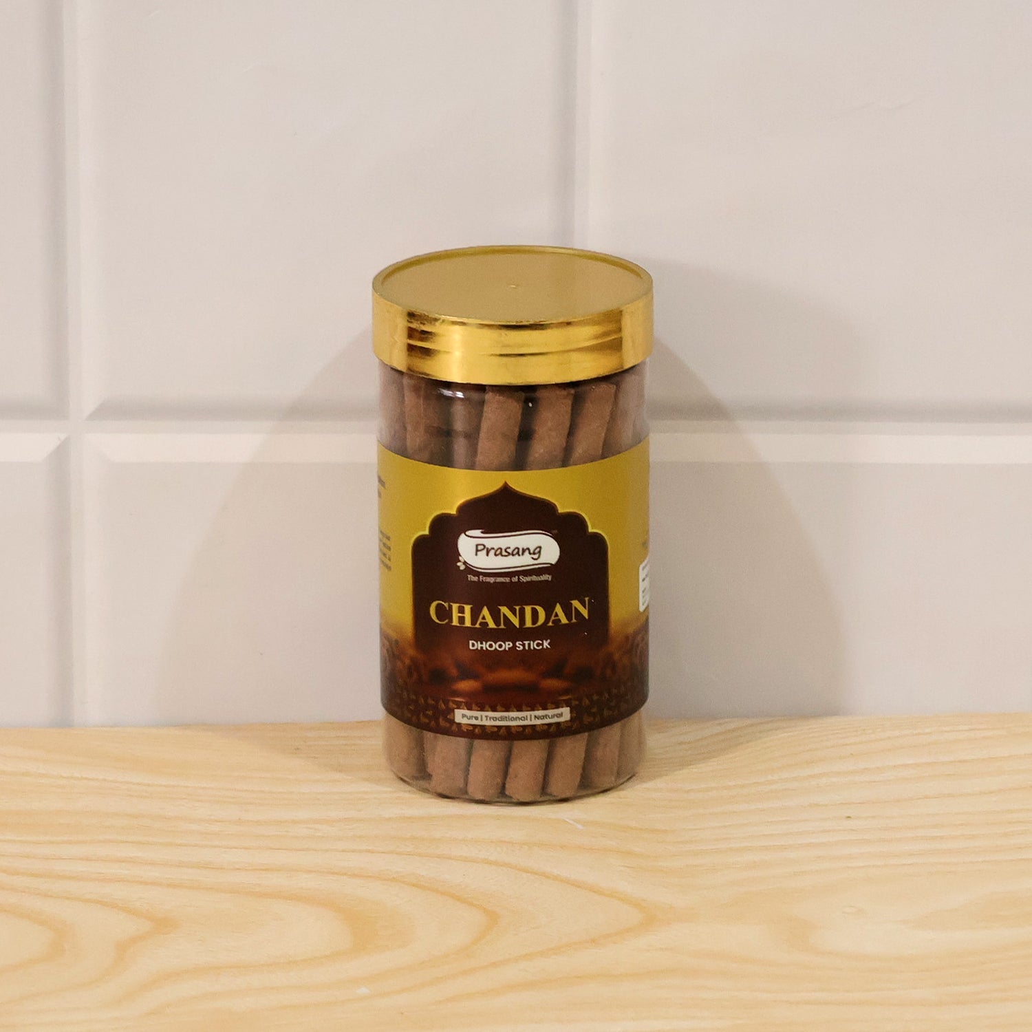 A clear jar of brown dhoop sticks with a gold lid rests on a light wooden surface against a tiled wall.