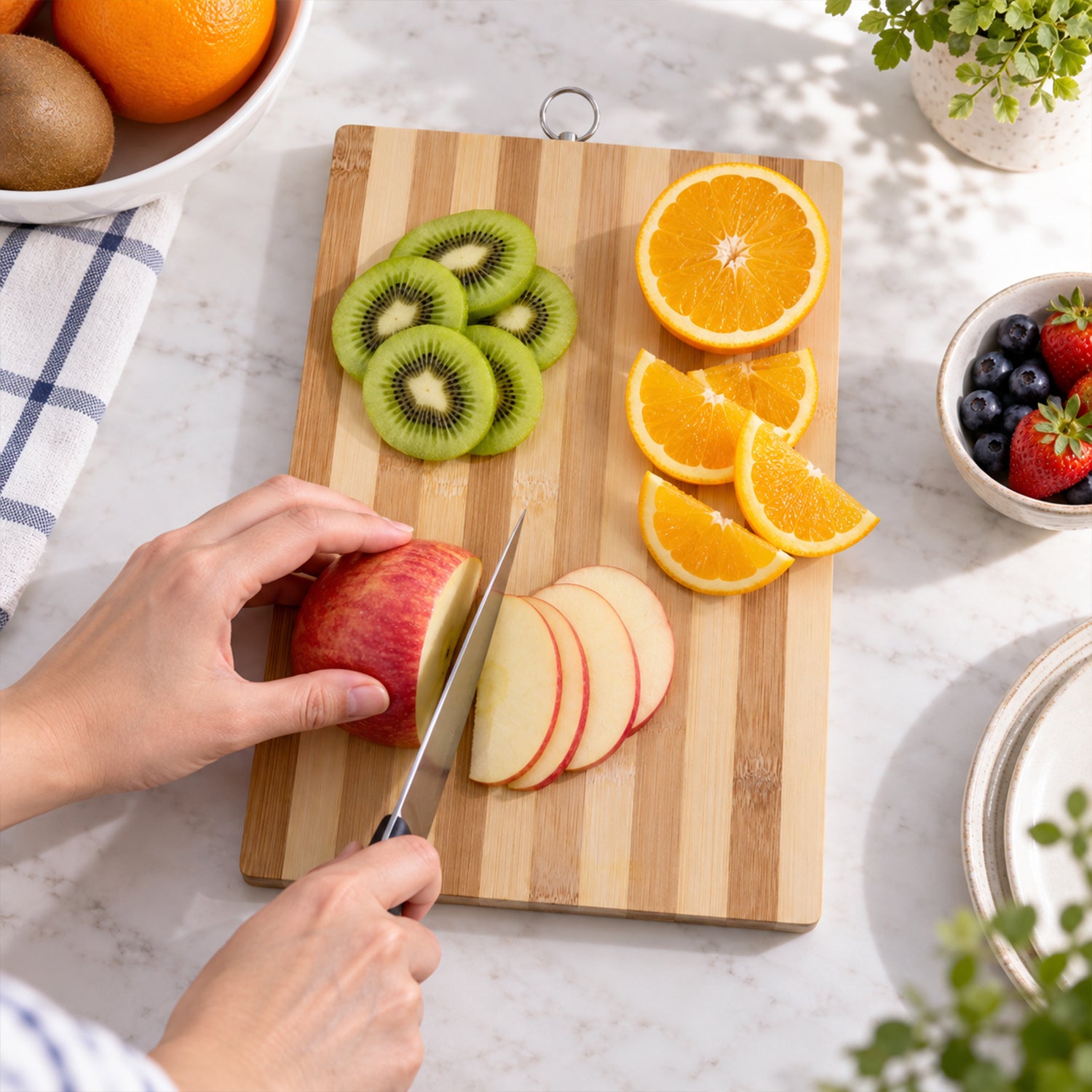 A person slices an apple on a striped bamboo board, surrounded by kiwi, orange, and berries on a white counter.