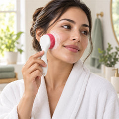 A woman in a white robe cleanses her cheek with a pink and white silicone face brush, creating foam.