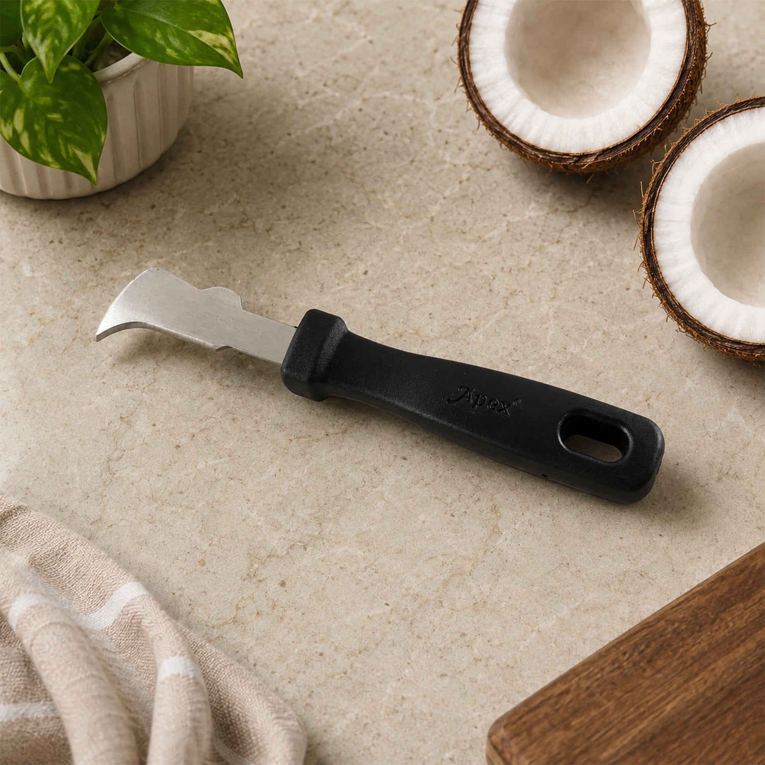 The Apex coconut meat removal tool rests on a light marble surface next to two coconut halves and a potted plant.