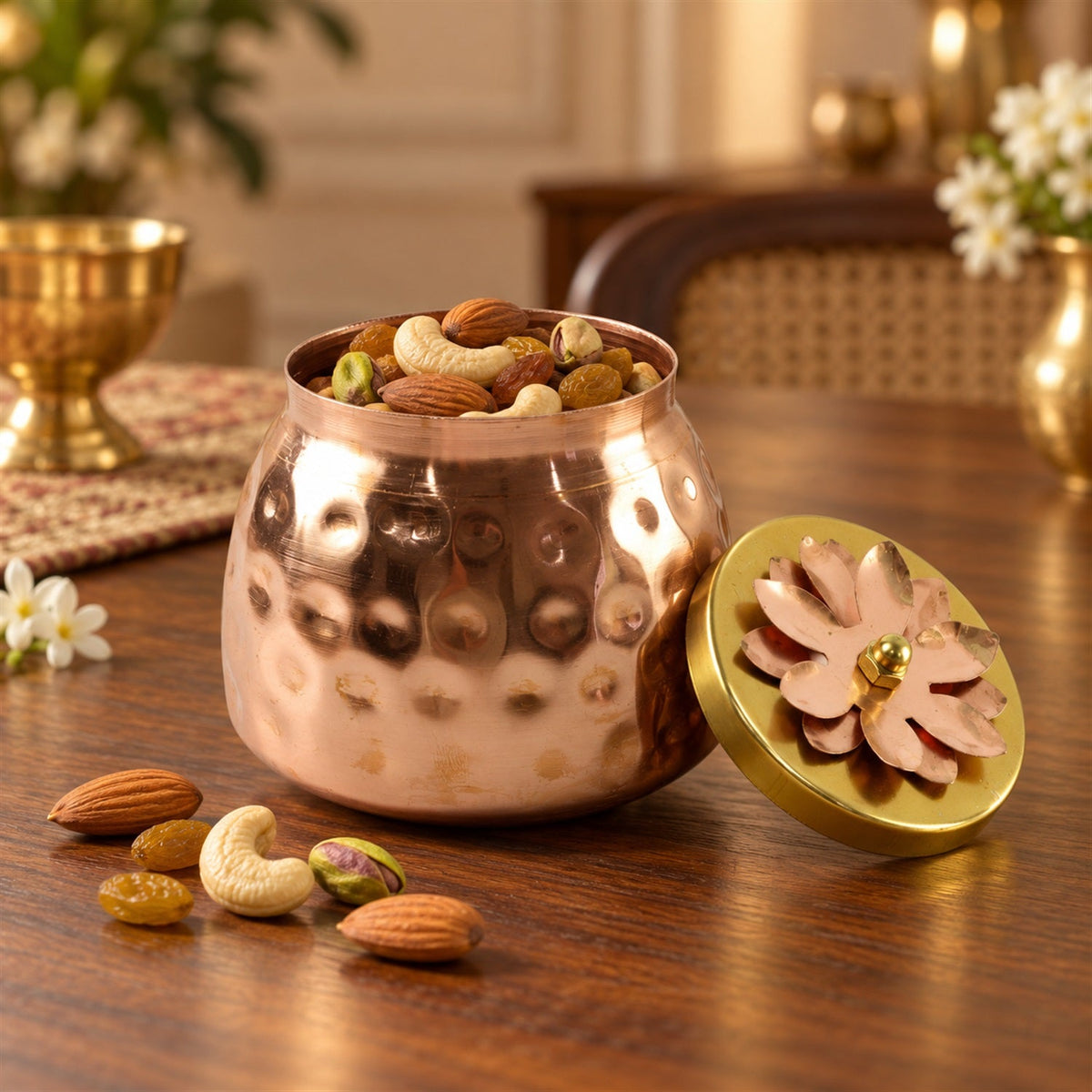 A hammered copper jar filled with mixed nuts, with a gold lid featuring a copper flower design resting beside it on a wooden