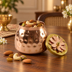 A hammered copper jar filled with mixed nuts, with a gold lid featuring a copper flower design resting beside it on a wooden
