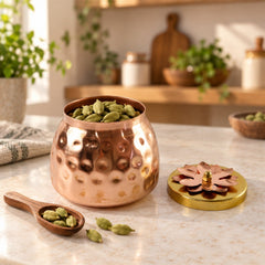 A hammered copper jar filled with green cardamom pods sits on a light countertop, with a wooden spoon holding more pods