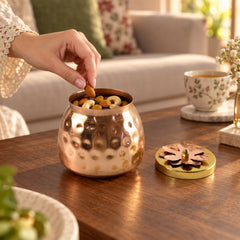 A hand reaches into a hammered copper jar filled with nuts, taking an almond, with the jar's decorative lid beside it on a