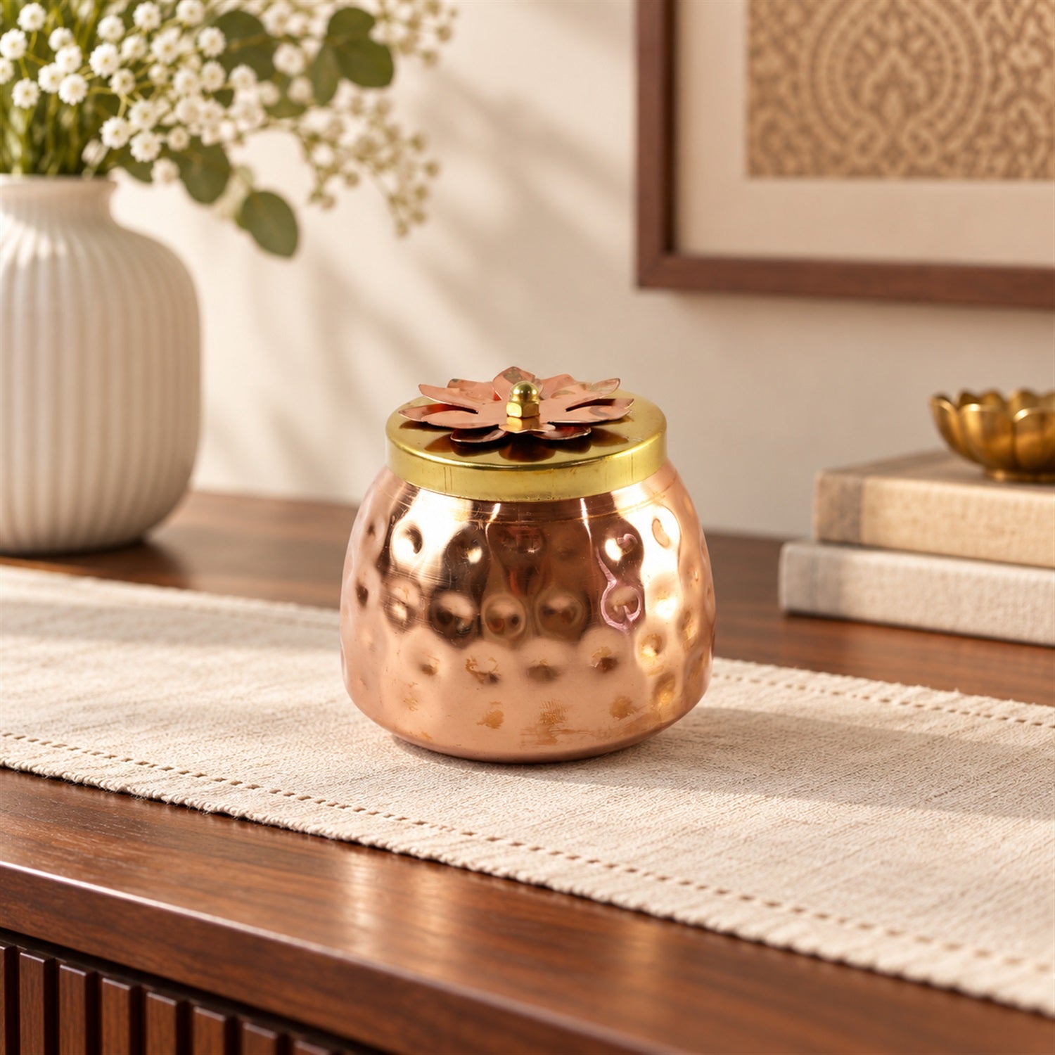 A hammered copper jar with a decorative flower lid sits on a light-colored table runner, on a dark wooden console with white