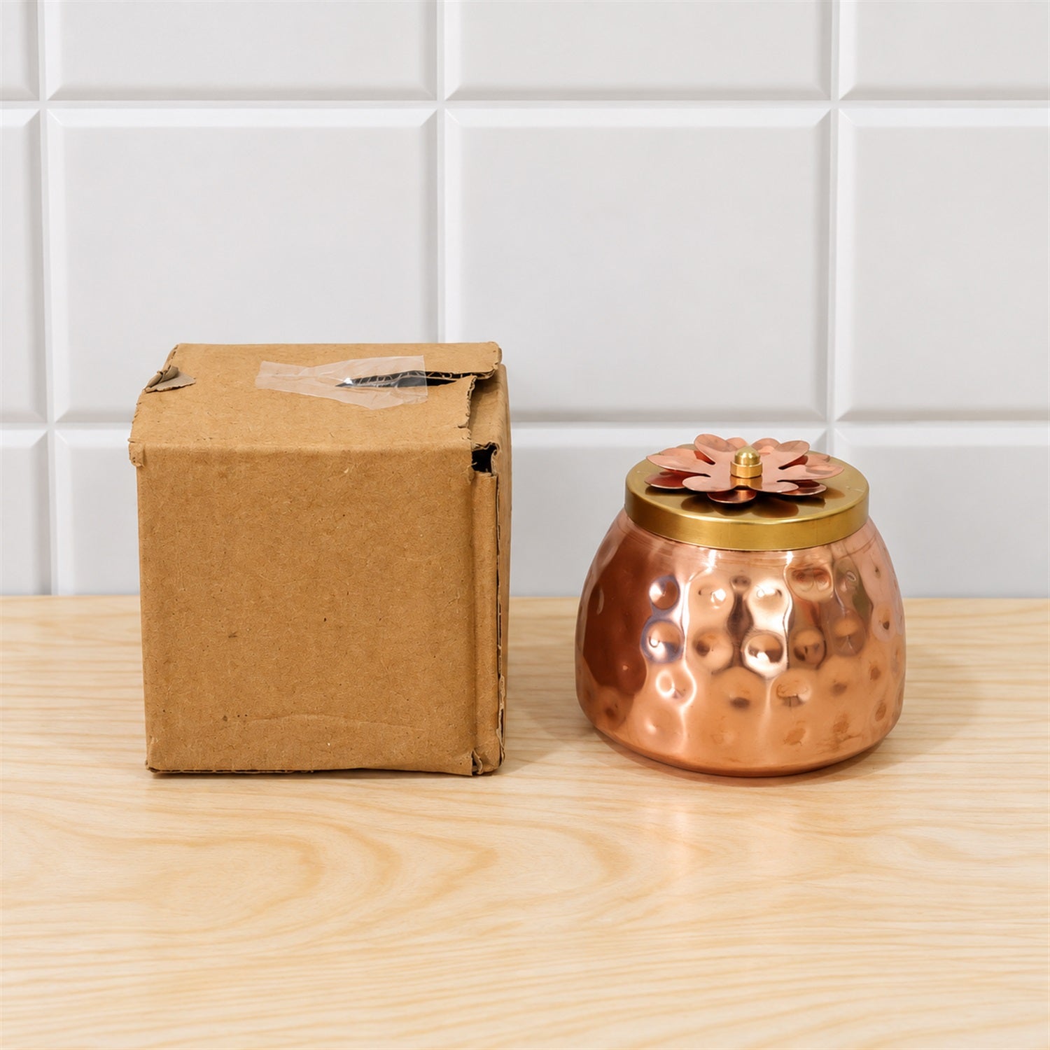 A small, square cardboard box sits next to a hammered copper jar with a decorative floral lid on a light wooden surface