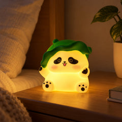 A glowing panda night light with a green leaf hat sits on a bedside table next to a plant.