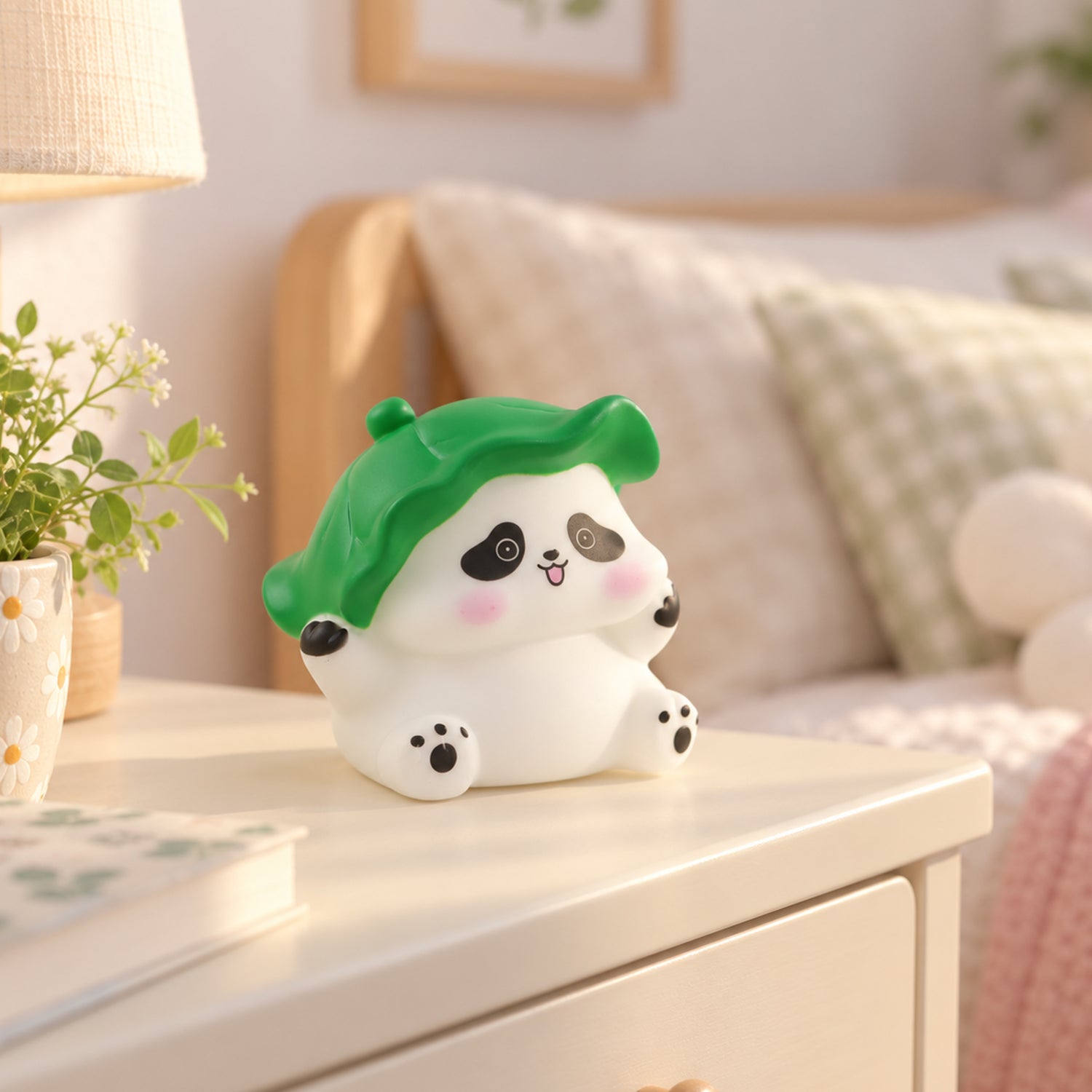 The panda night light, unlit, sits on a light wooden bedside table in a bright bedroom with a plant and lamp.