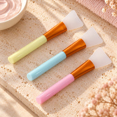 Three silicone face mask brushes with pastel handles in yellow, blue, and pink, on a textured stone surface with a bowl of