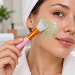 A woman applies a green face mask to her cheek with a pink-handled silicone brush, wearing a white robe and head towel.