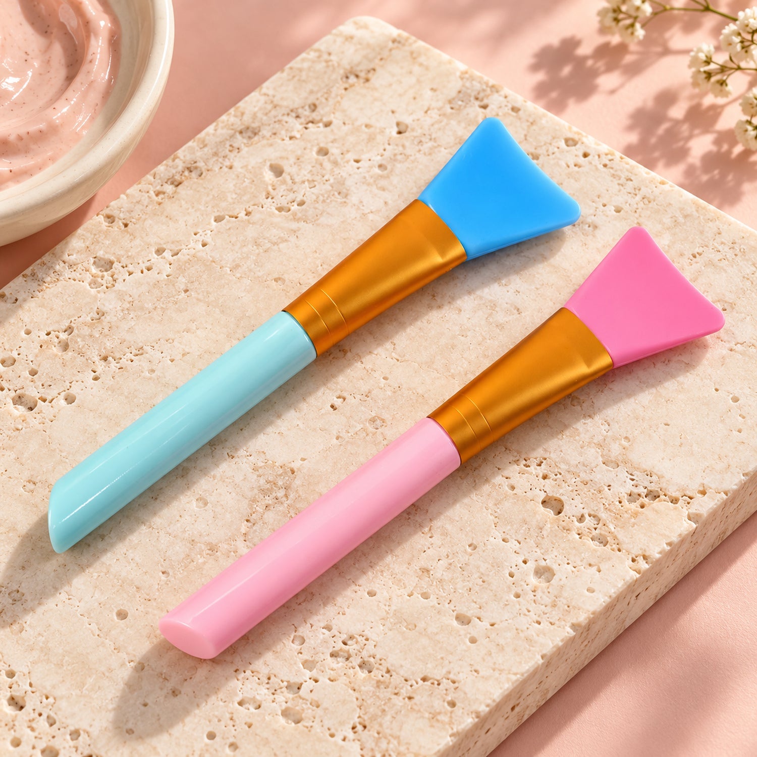 Two silicone face mask brushes, one pink and one blue, rest on a light marble surface.