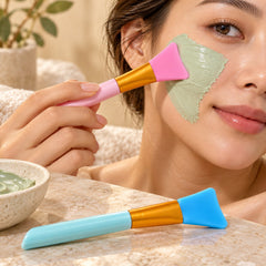 A woman applies a green face mask with a pink silicone brush, while a blue brush rests on a marble surface.