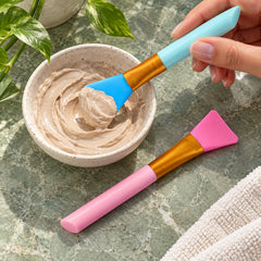 A hand uses a blue silicone brush to scoop a brown face mask from a ceramic bowl, with a pink brush alongside.