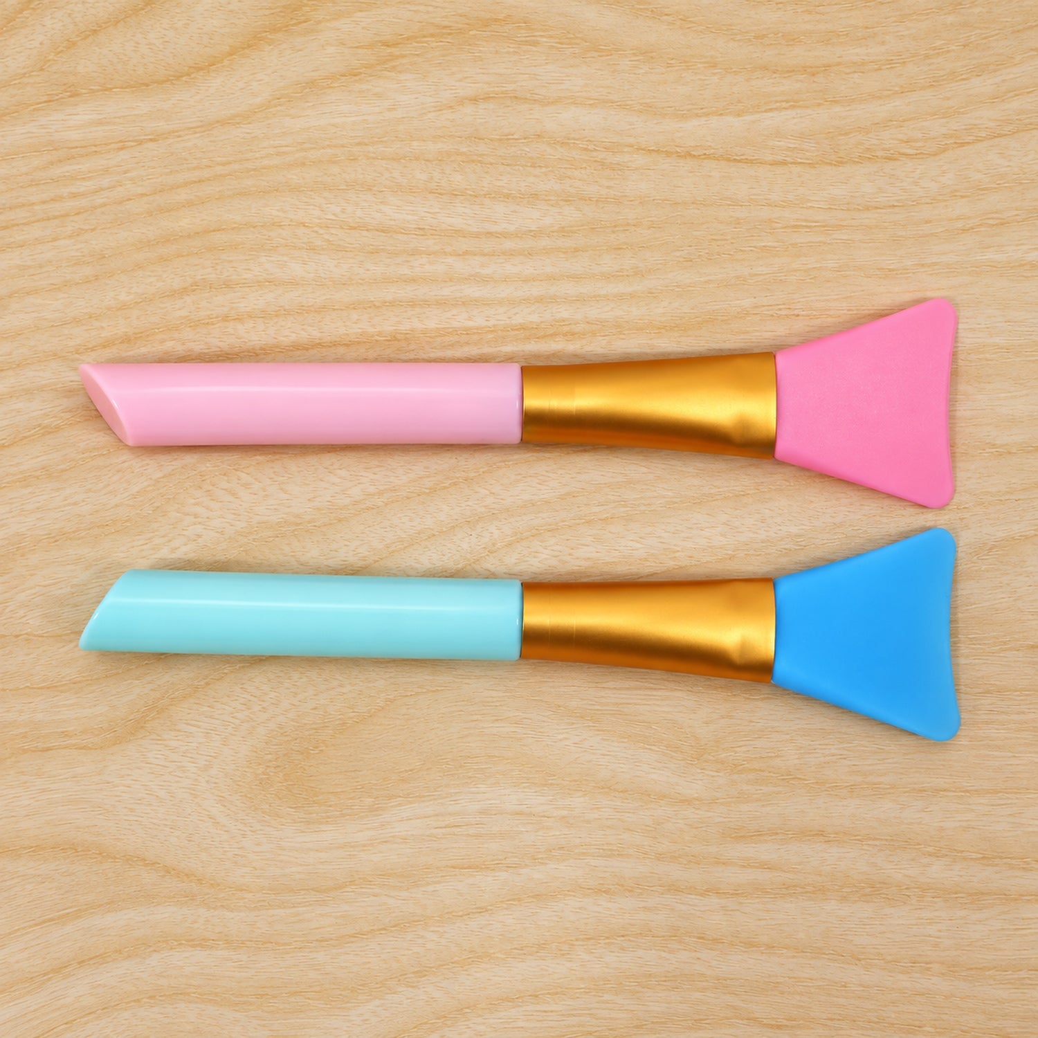 A pink and a blue silicone face mask brush lie horizontally on a light wooden surface.
