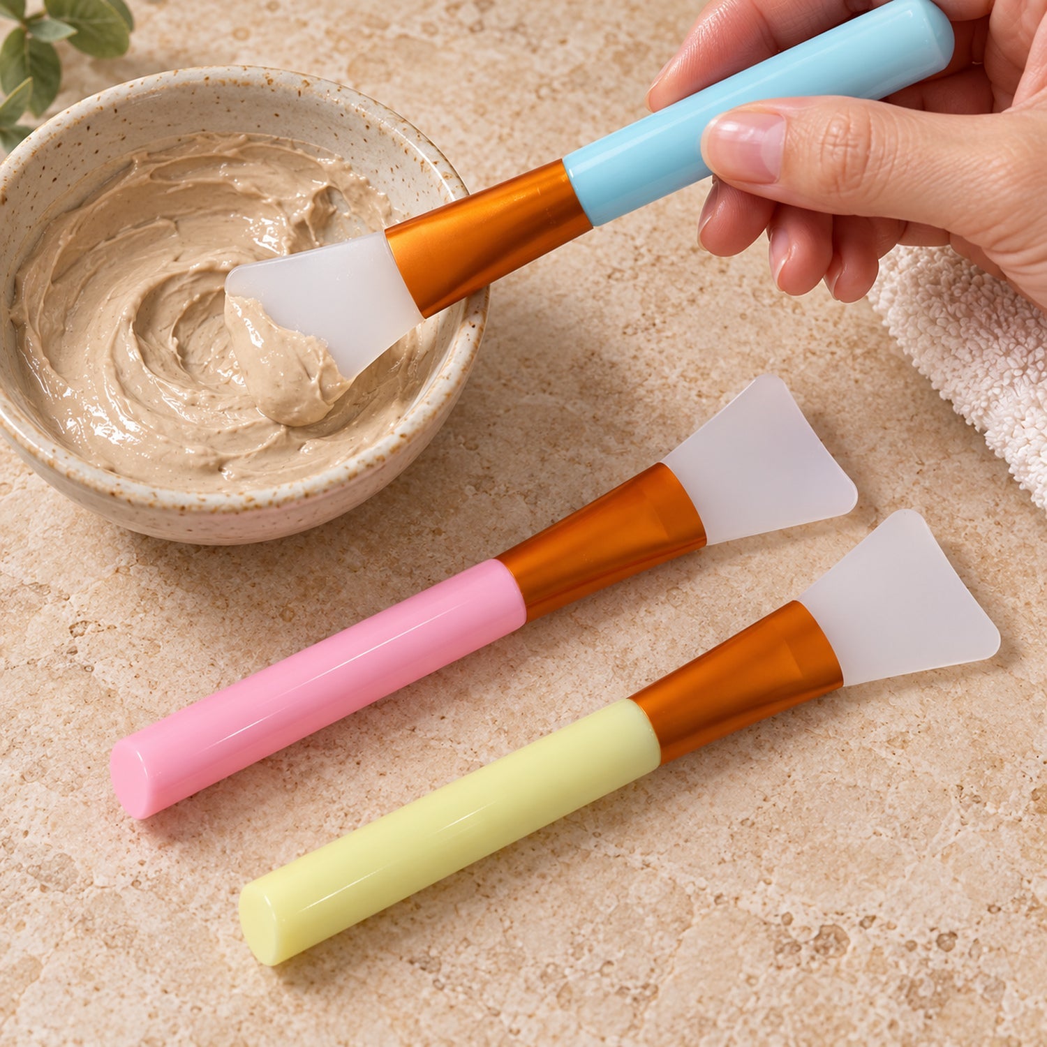 A hand dips a blue-handled silicone brush into a bowl of brown face mask, with pink and yellow brushes lying beside it.