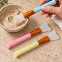 A hand dips a blue-handled silicone brush into a bowl of brown face mask, with pink and yellow brushes lying beside it.
