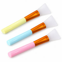 Three silicone face mask brushes with pastel handles in yellow, blue, and pink, arranged diagonally on a white background.