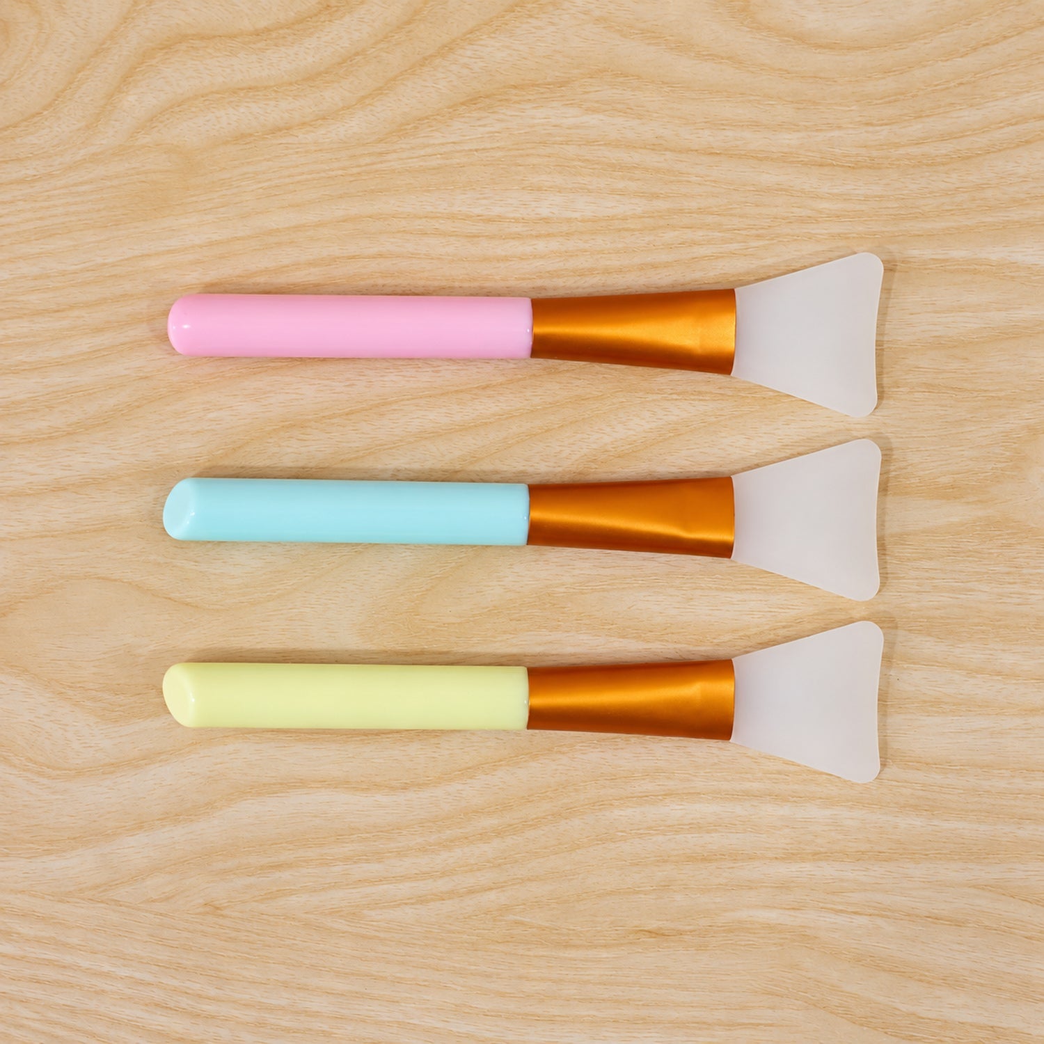 Three silicone face mask brushes with pastel handles in pink, blue, and yellow, arranged horizontally on a light wooden