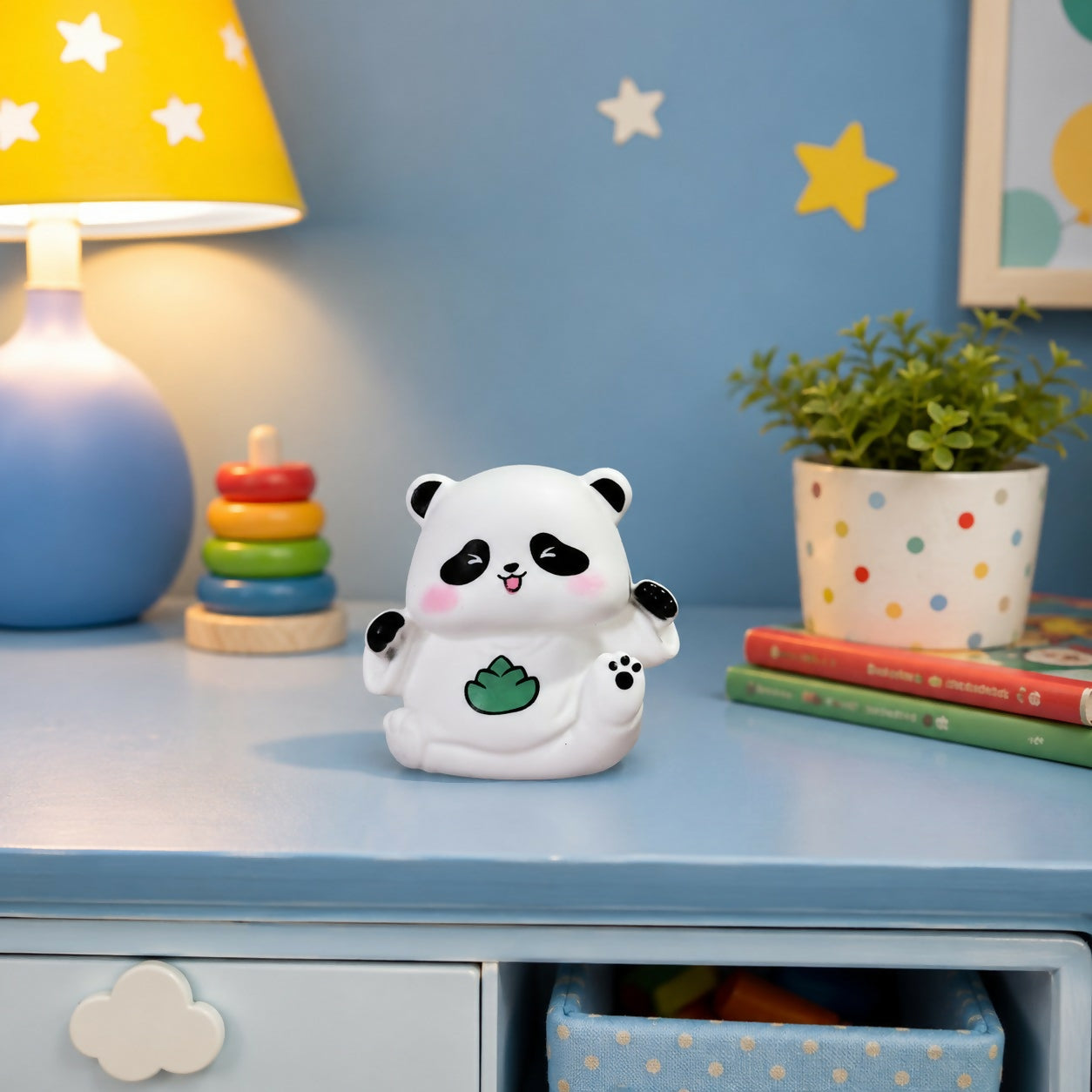 A white panda night light with black ears and eye patches, pink cheeks, and a green leaf design sits on a blue table.