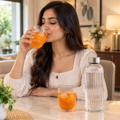 A woman in a light-colored shirt sips from a ribbed glass tumbler filled with an orange beverage and ice, with a matching