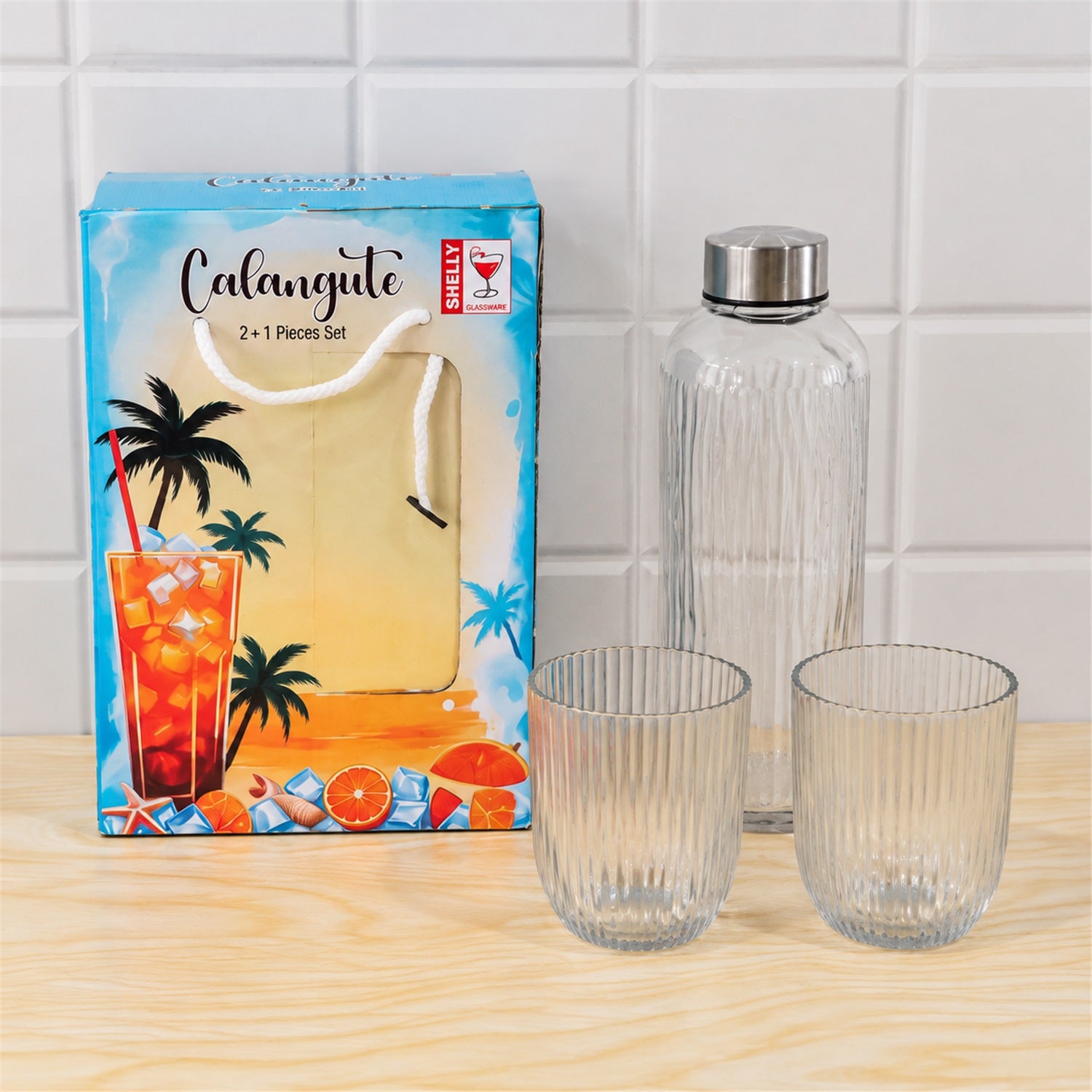 A blue gift box with a tropical design is placed next to an empty clear ribbed glass bottle and two empty matching tumblers
