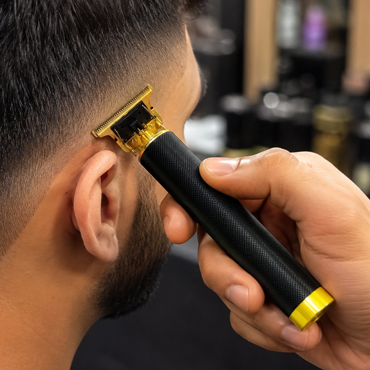A close-up shows a man's ear and the side of his head as a hand uses a black and gold trimmer to cut his hair.