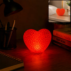 A red heart-shaped LED night lamp illuminates a dark desk next to a black desk lamp and books.