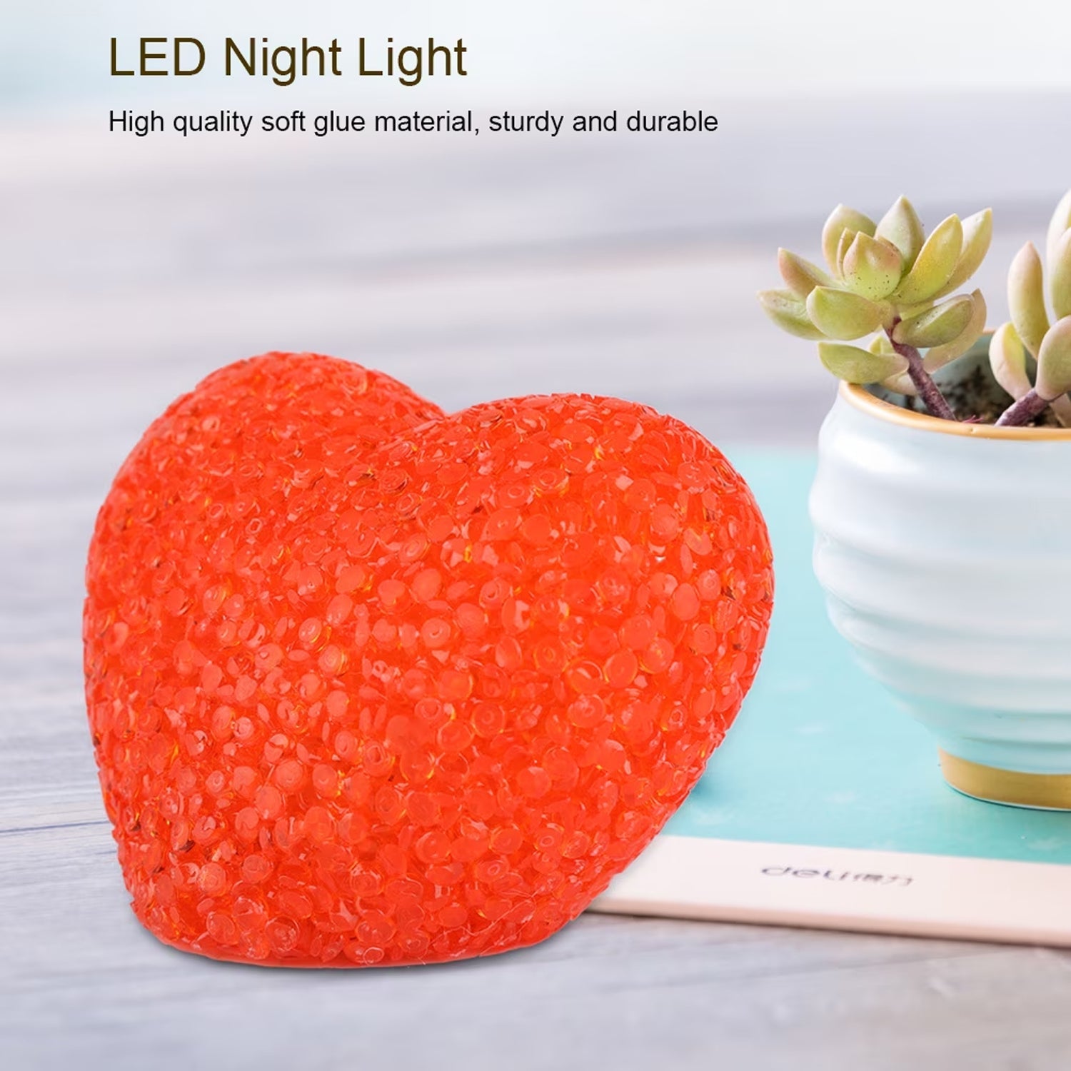 A textured orange heart-shaped LED night light sits on a light blue surface next to a potted succulent.