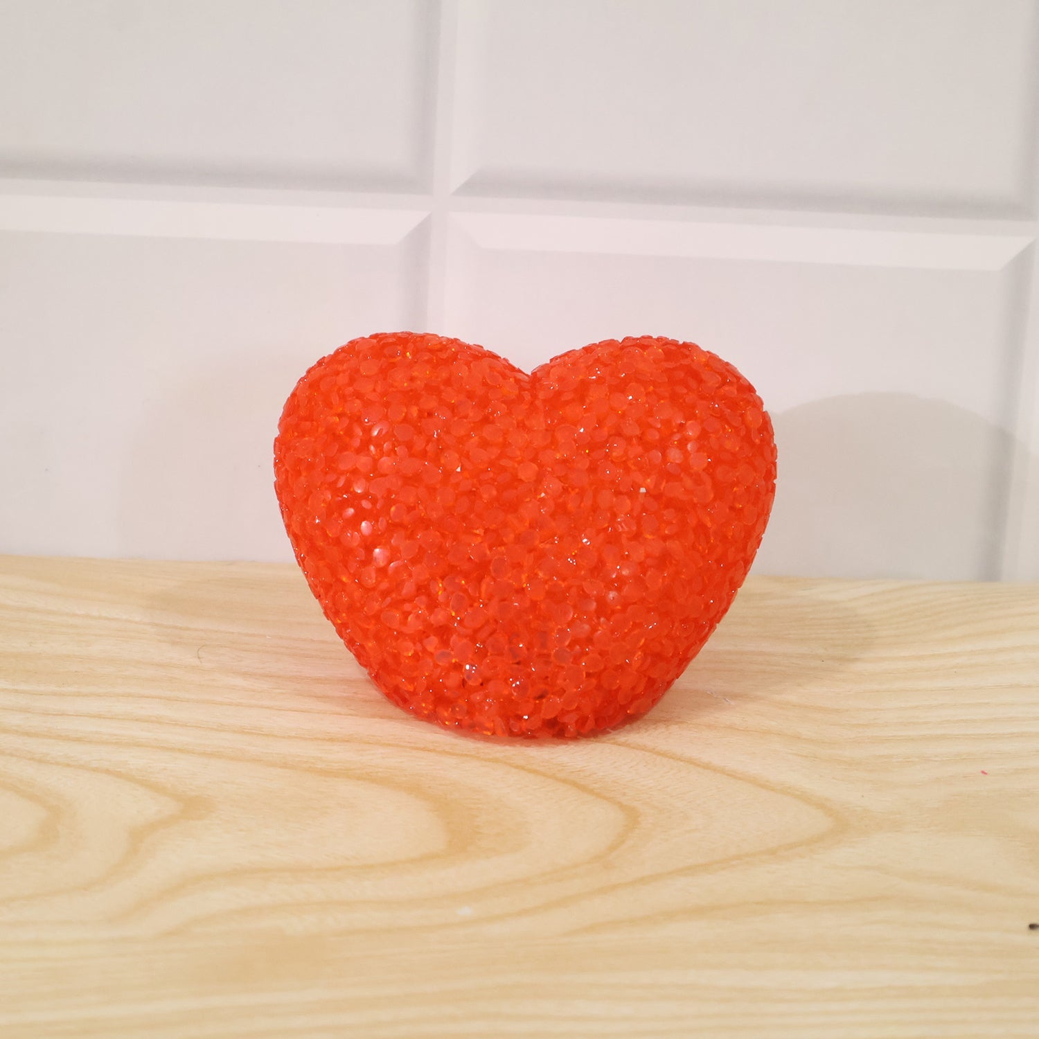 A textured orange heart-shaped LED lamp is centered on a light wooden surface against a white tiled wall.