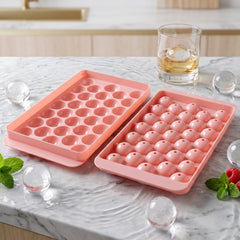 Two pink plastic ice ball trays on a marble counter, one filled with ice balls, next to a whiskey glass.