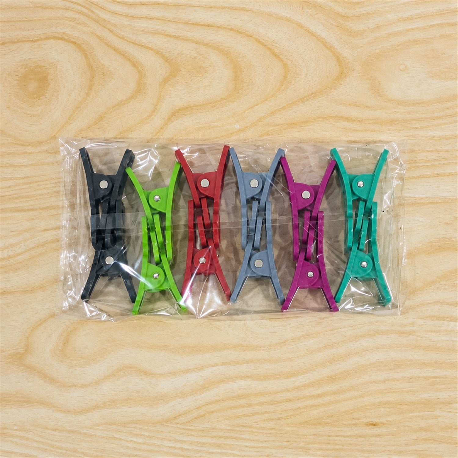 Six pairs of colorful plastic clothes clips, still in their clear plastic packaging, are displayed on a light wooden surface.