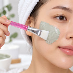 A woman with a white towel on her head applies a green face mask to her cheek with a pink-handled brush.