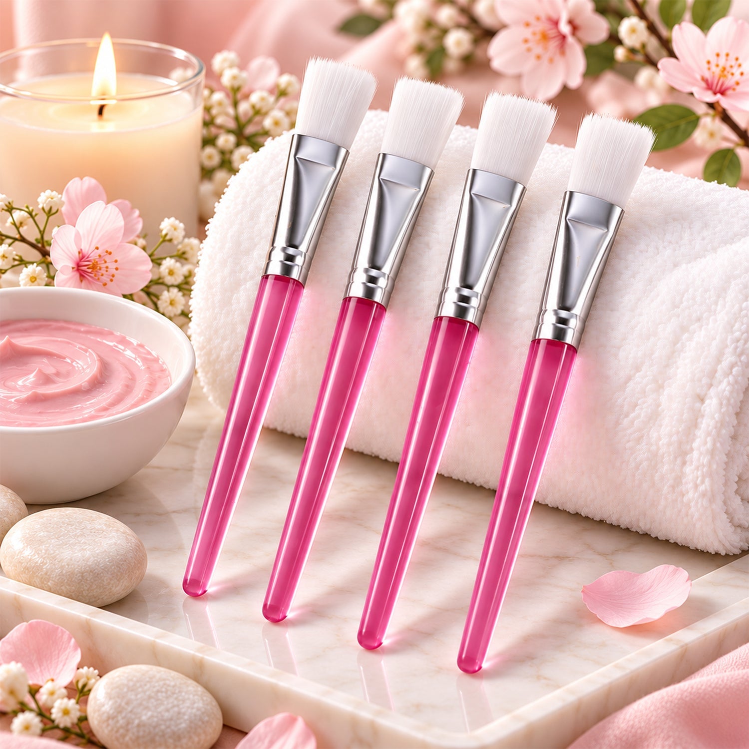 Four pink-handled face mask brushes rest on a white towel next to a lit candle, pink mask, and decorative stones.