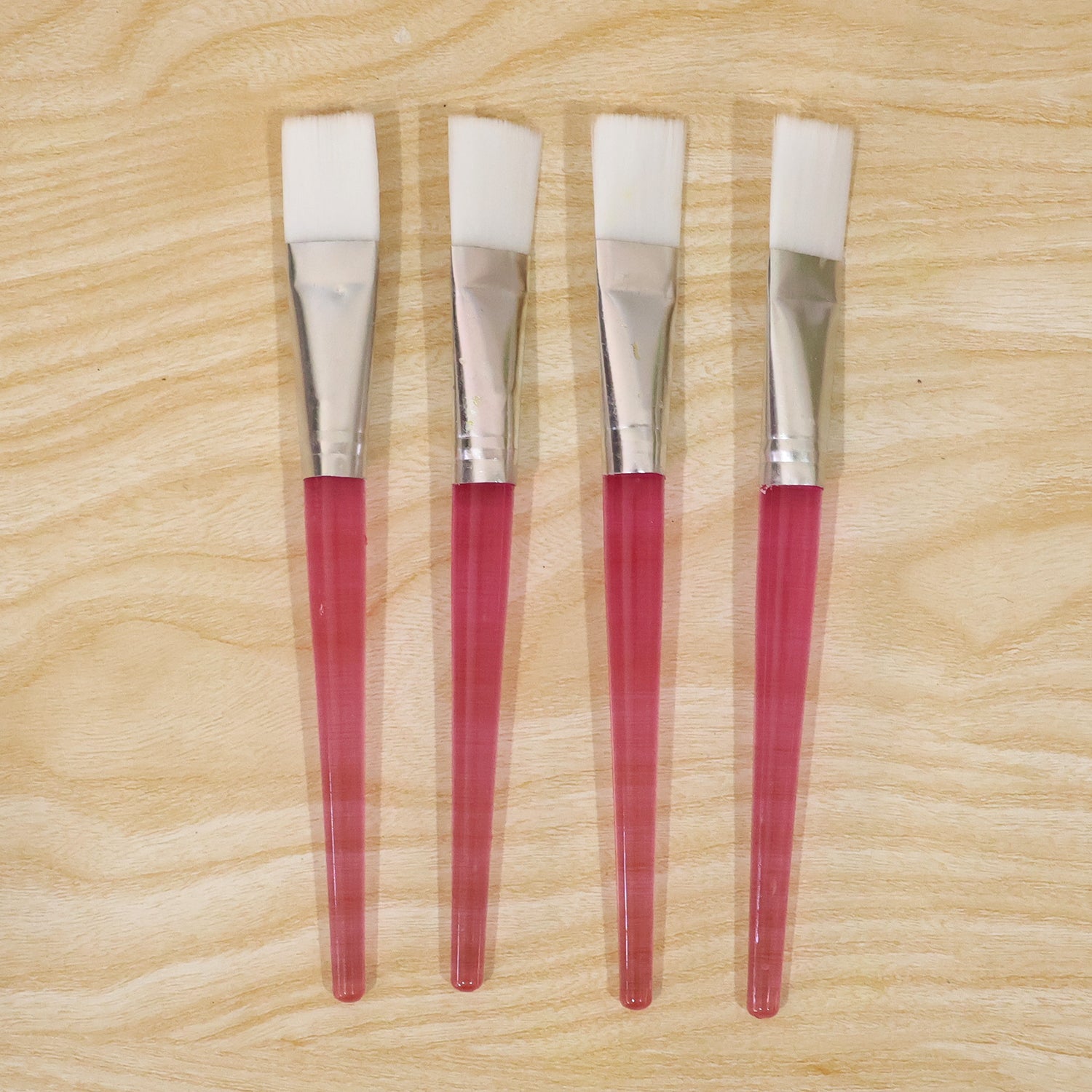Four pink-handled face mask brushes with white bristles are neatly arranged on a light wooden surface.