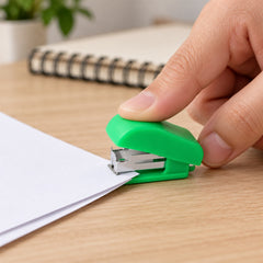 A hand presses down on a small green stapler, joining two sheets of white paper on a light wooden table.