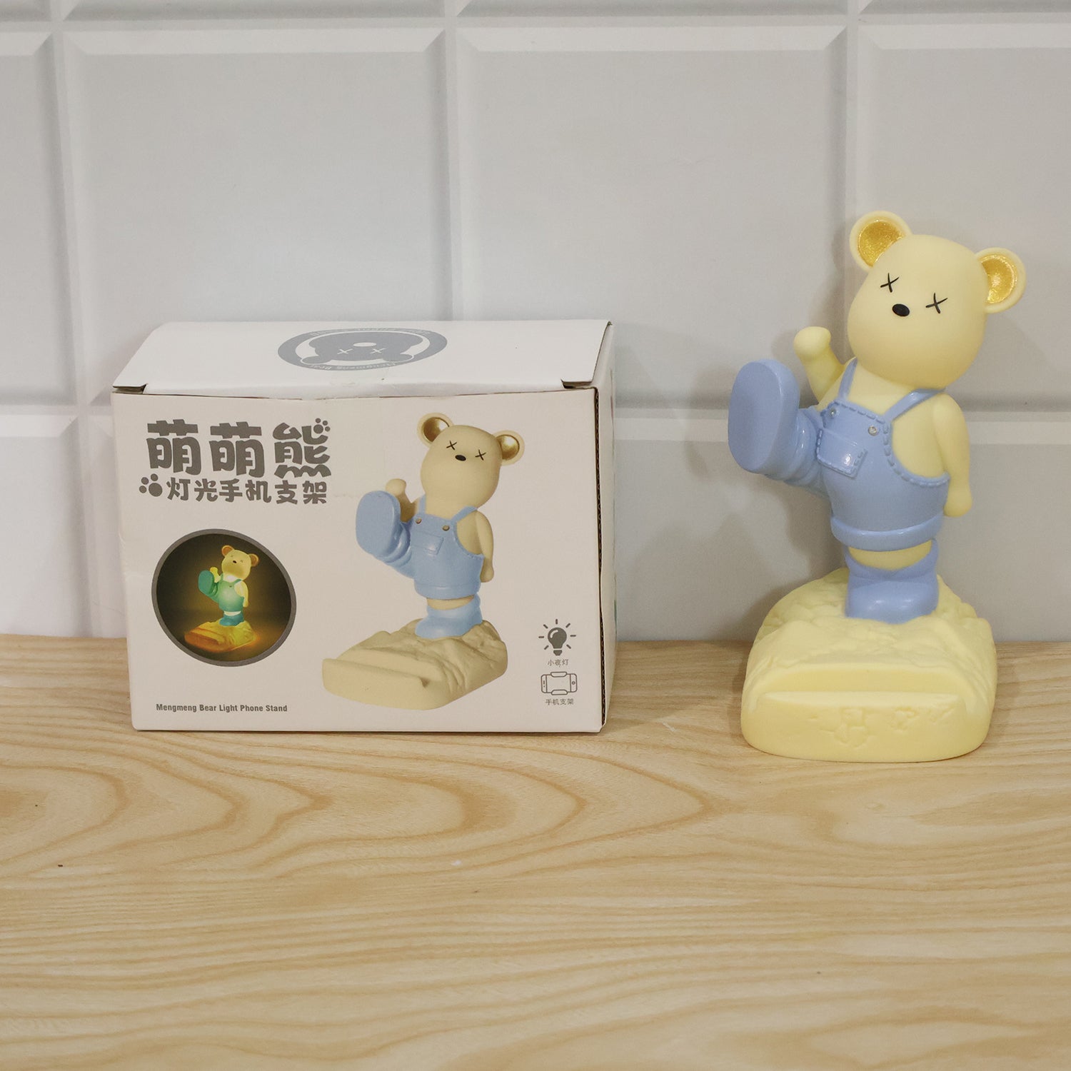 A light yellow bear phone stand next to its white product box, both on a light wooden surface.