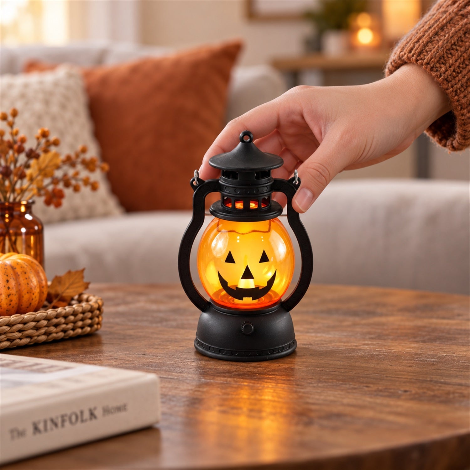 A hand gently touches a glowing pumpkin lantern on a wooden coffee table, surrounded by autumn decor.