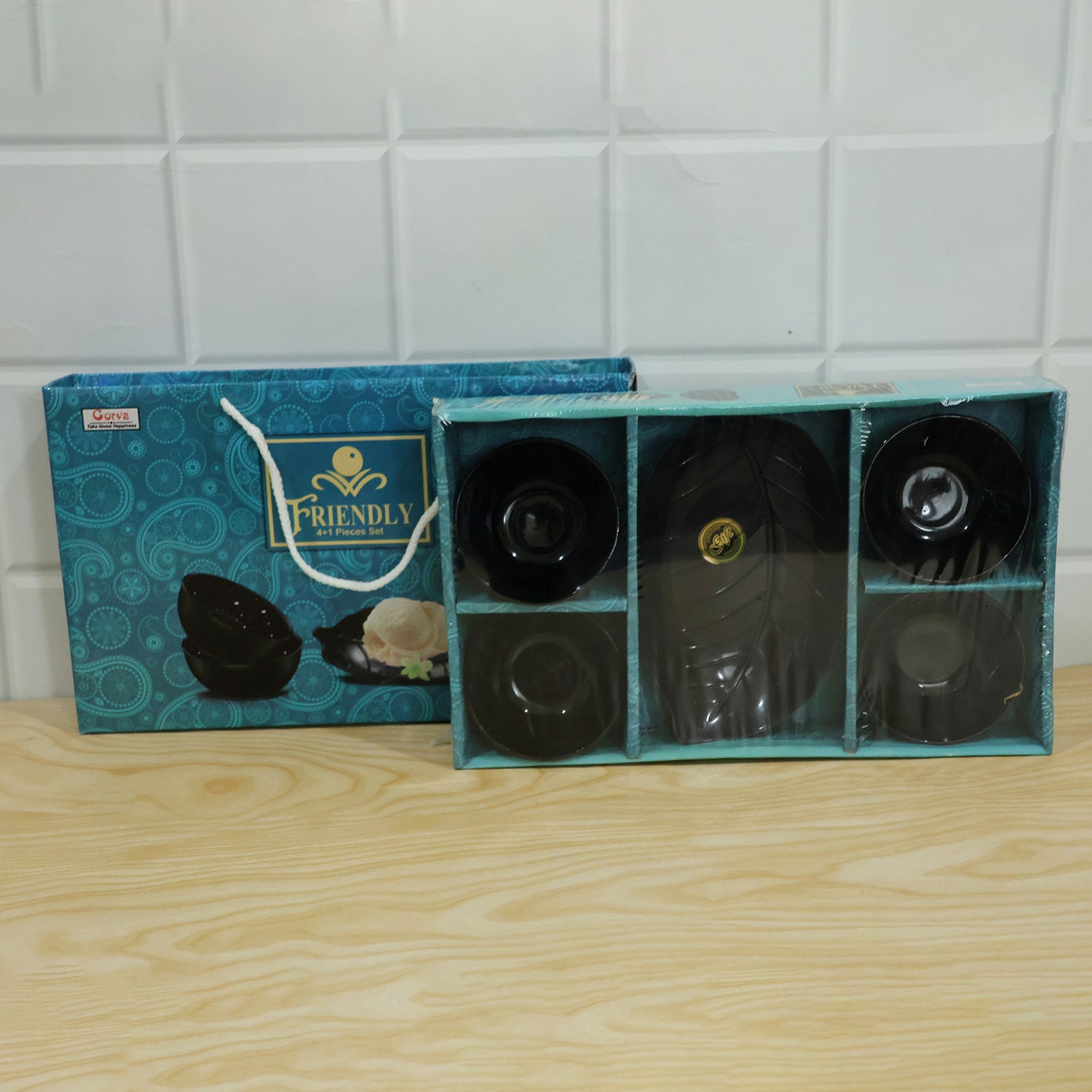 A blue patterned gift box with a black leaf-shaped platter and four bowls is on a light wooden surface.
