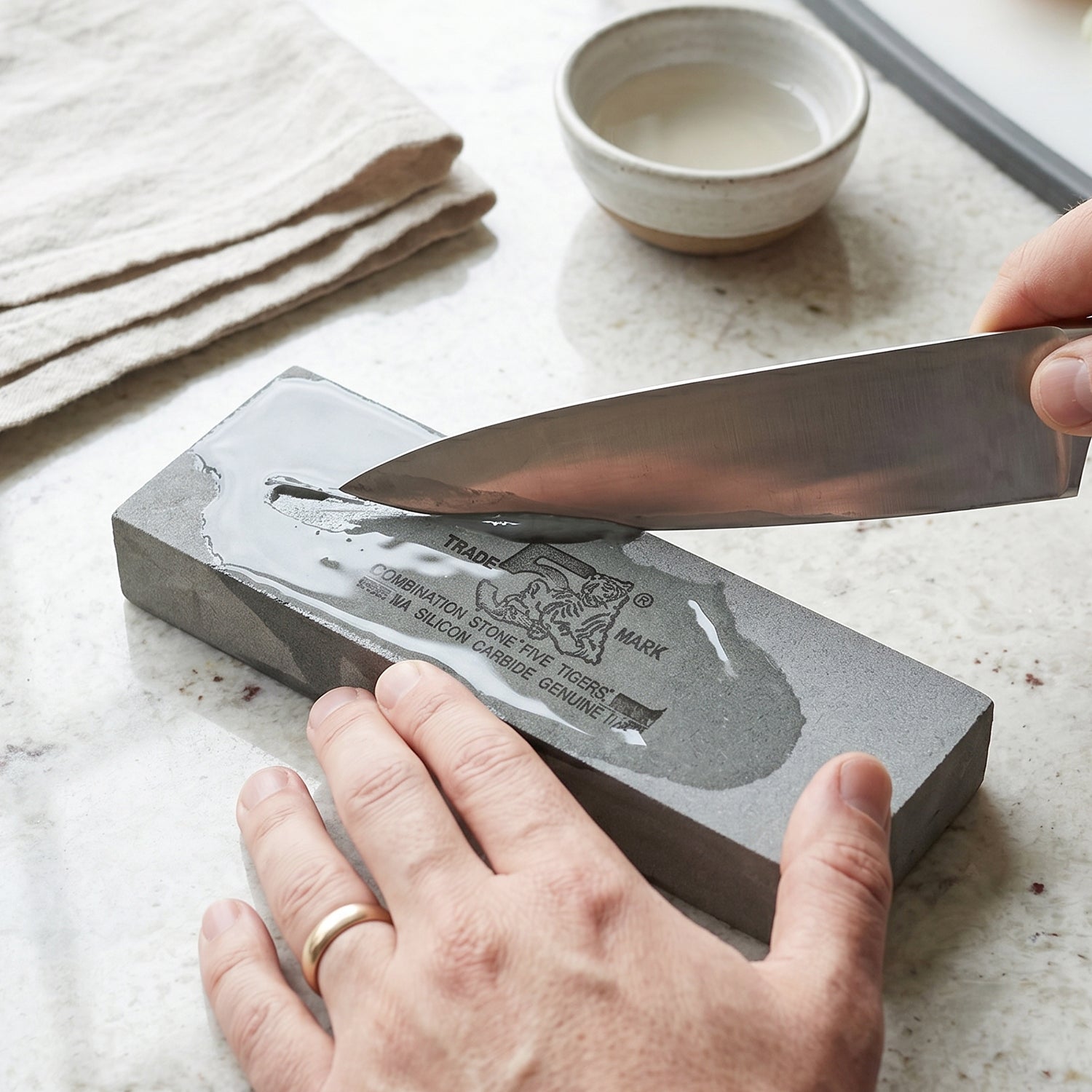 Hands sharpen a knife on a wet gray silicon carbide whetstone, with a bowl of water nearby.