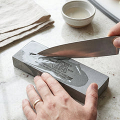 Hands sharpen a knife on a wet gray silicon carbide whetstone, with a bowl of water nearby.