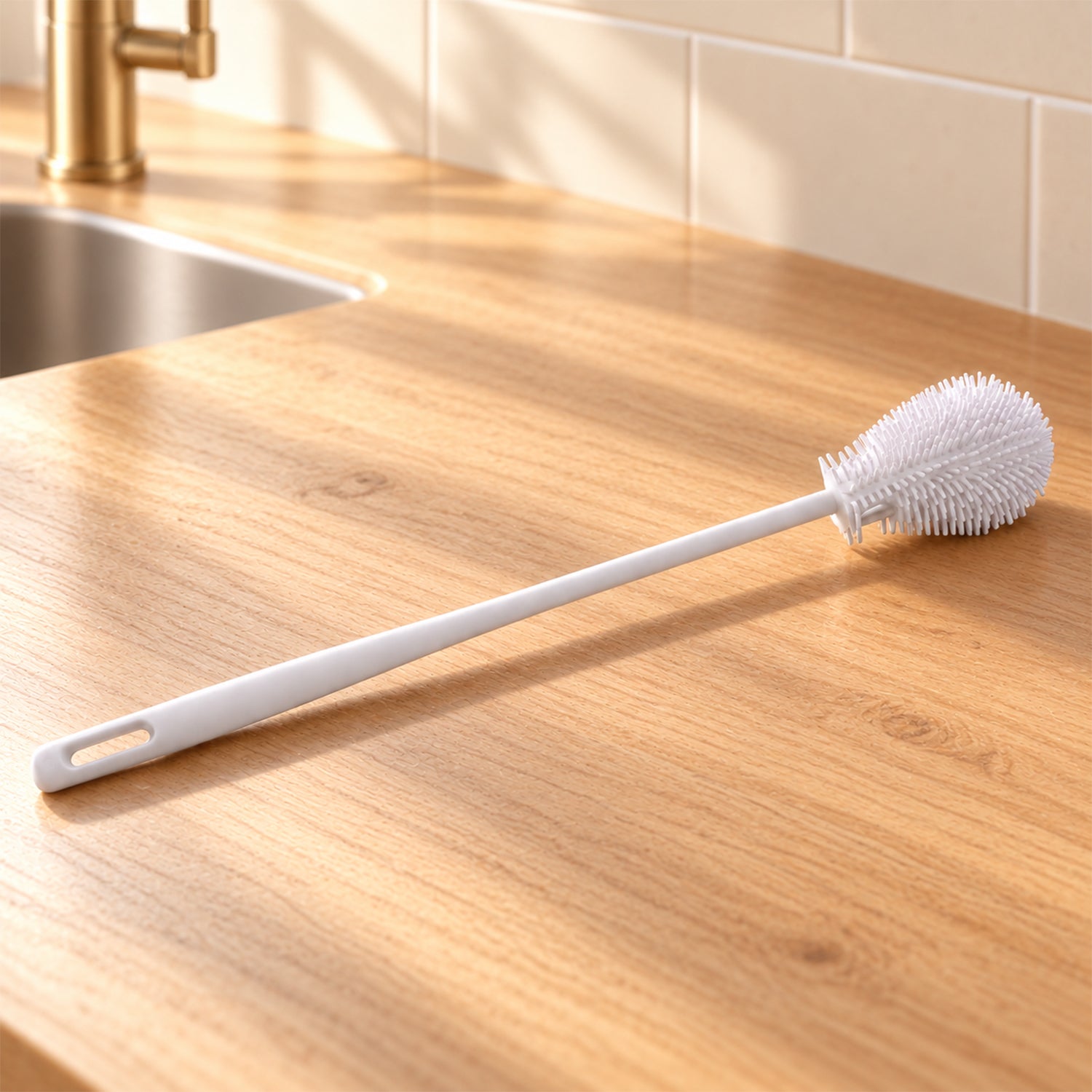 A white silicone bottle cleaning brush rests on a light wooden countertop next to a kitchen sink with a gold faucet.