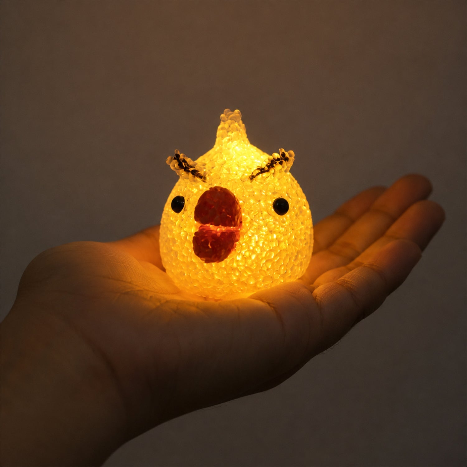 A glowing yellow chick-shaped night lamp with a red beak and black eyes held in the palm of a hand.